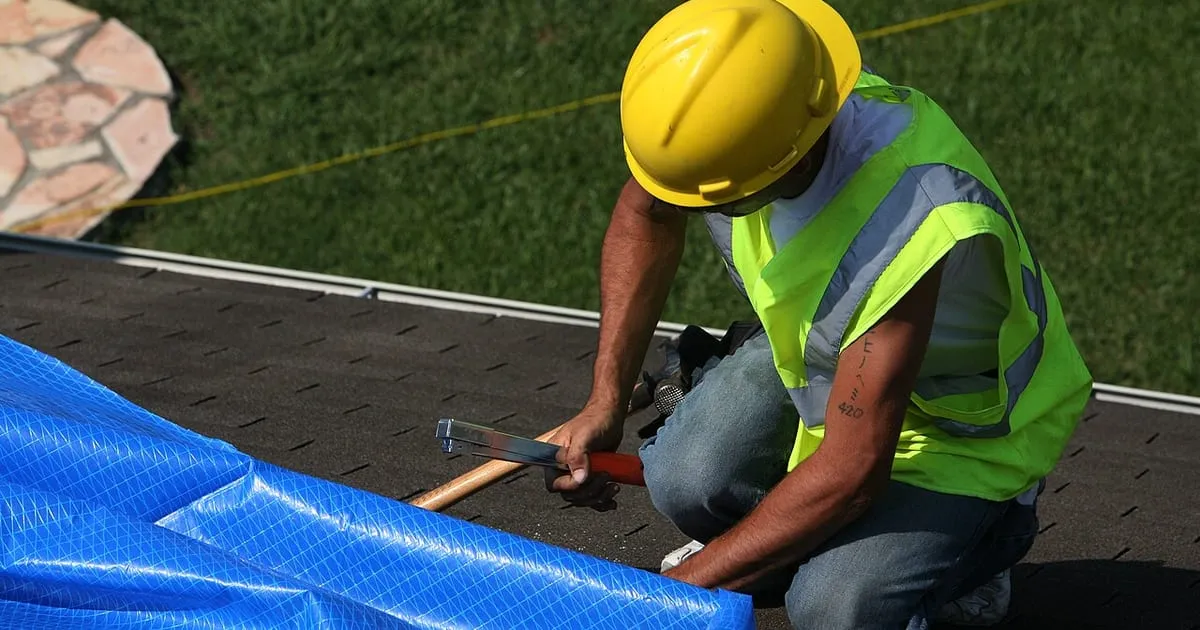 A contractor in a yellow hard hat and high-visibility vest kneeling on a roof and fixing a blue tarp in place