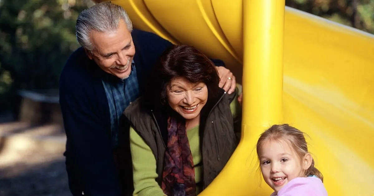 Smiling grandparents leaning over a yellow playground slide where their young granddaughter sits