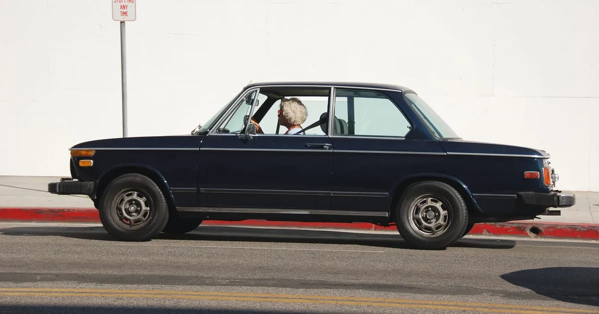 An older dark blue two-door car being driven along a city street past a white wall