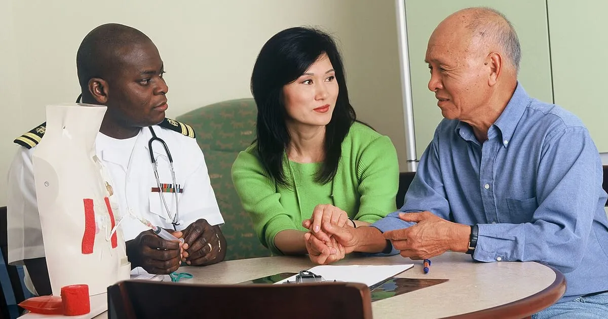 An older man and a family member holding hands at a table while talking with a doctor