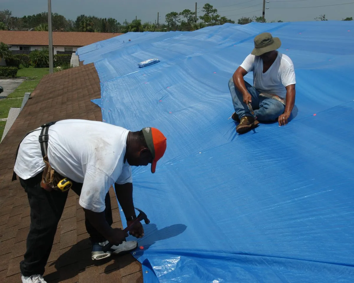 Workers nailing a blue tarp over a storm-damaged roof in Florida